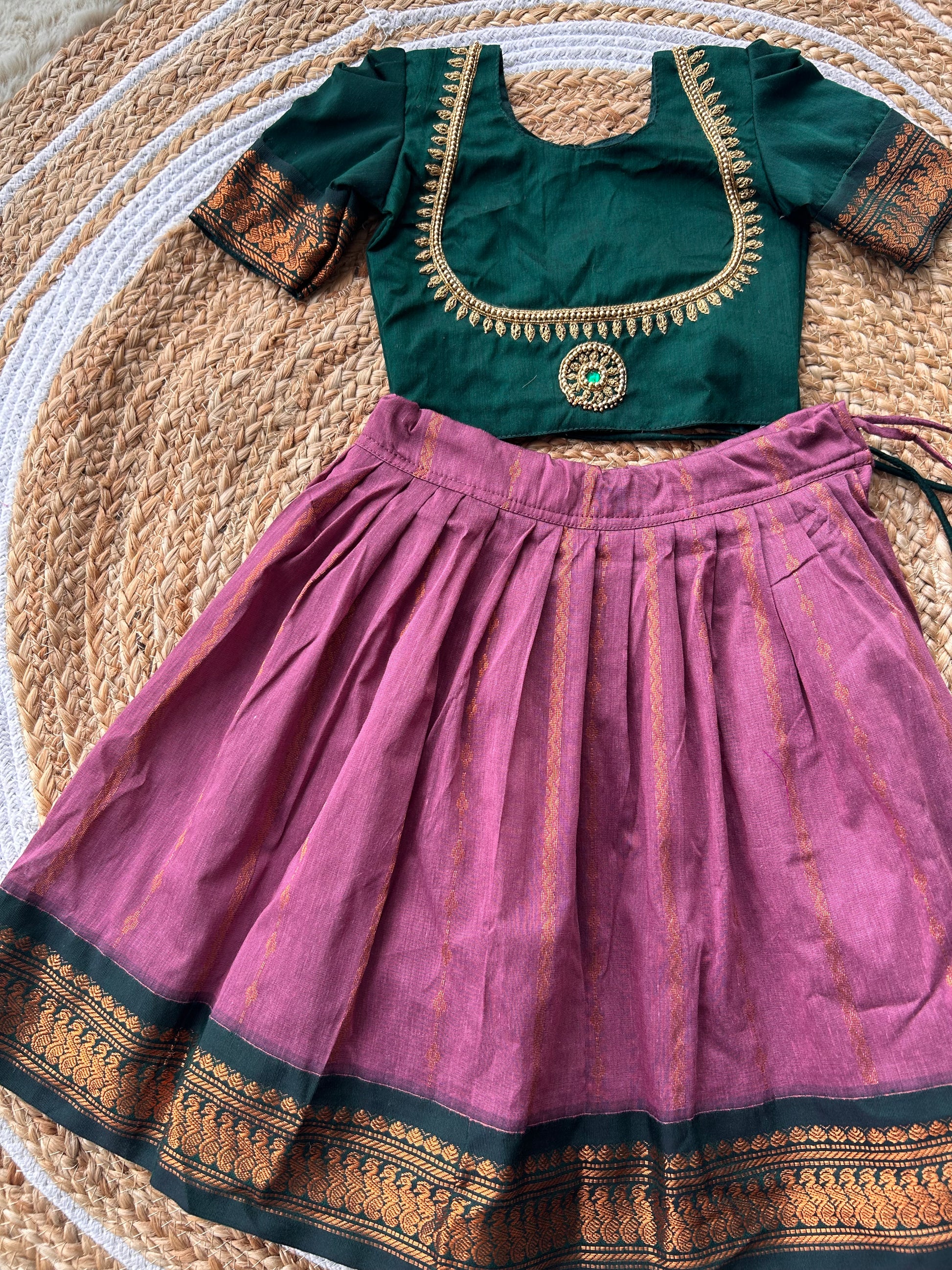 A traditional Indian ethnic wear set consisting of a dusty purple pleated skirt and a dark green embroidered blouse, laid flat on a woven straw mat.